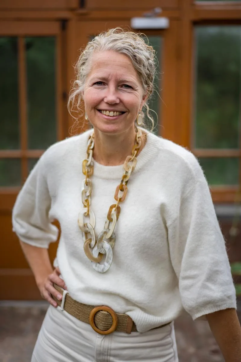 Margje Kooiker staand, glimlachend voor een houten gevel